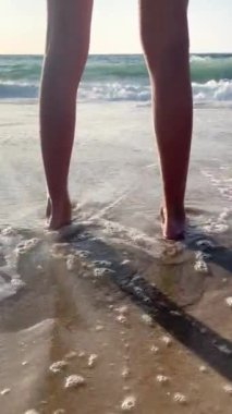 Okyanus suyunda ayakta duran çocuk bacaklarına yakından bak. Atlantik Okyanusu kıyısında duran kız. Çıplak kadın bacakları ve plajları yıkayan dalgaların video görüntüsünü kapat..