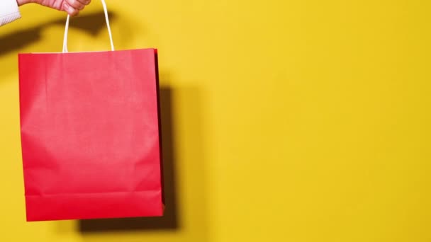 ショッピングや販売のコンセプトのフルHdビデオ 女性の手は明るい黄色の背景で隔離された赤い買い物用紙袋を保持しています コピースペースでビデオバナーを販売 — ストック動画