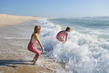 Two young girls are playing in the ocean waves on a sandy beach, laughing and splashing in the foamy water. The sun is shining brightly, creating a fun and carefree summer scene clipart