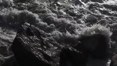 Dramatik okyanus dalgalarının karanlık kıyı kayalarına çarpıp sıçrama ve köpüklü su oluşturduğu bir video. Güç, esneklik ve deniz manzarası teması için ideal bir doğa sahnesi..