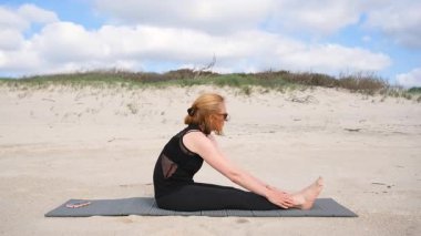 Kumlu yüzeyde yoga yapan bir kadının videosu, minder üzerinde öne eğilip poz verirken, açık bir gökyüzü altında doğal çevrede rahatlığı ve esnekliği kucaklarken.