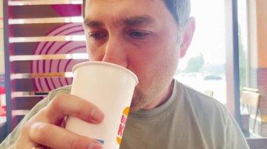 Burger King bardağından içen bir adamın yakın çekimi. Rahatlamış ve düşünceli görünüyor. Parlak, modern bir restoran ortamında ferahlatıcı bir içeceğin tadını çıkarıyor.