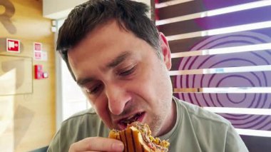 Bir adam fast-food restoranında ızgara burgerden büyük bir ısırık alıyor. Rahat bir atmosferde sıradan bir yemekten hoşlanır..