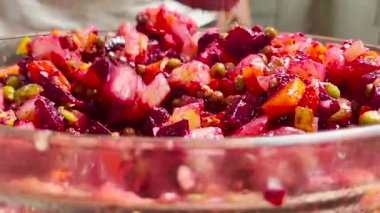 Canlı sebze salatasıyla cam kasede kaşığı karıştıran bir elin yakın çekimi. Renkli malzemeler arasında doğal mutfak ışığında pancar, havuç, bezelye ve patates yer alıyor.