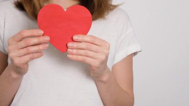 Beyaz tişörtlü genç bir kadın gökkuşağı renklerine boyanmış bir kalp taşıyor. LGBTQ gururunu, sevgiyi, dahil edilmeyi ve kimliğini simgeleyen güçlü bir görsel kavram..