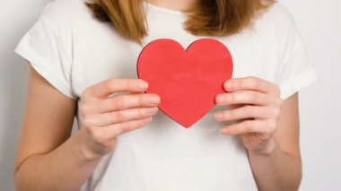 Beyaz tişörtlü genç bir kadın elinde kırmızı bir kalp sembolü tutuyor. Sevgi, ilgi, romantizm ve insan ilişkilerini temsil eden minimalist ve duygusal bir kavram.