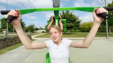 İki neşeli kız, muhtemelen arkadaşları ya da kız kardeşleri, parktaki spor aletlerinde birlikte oynuyorlar. Halka açık bir spor sahasında gülerler ve güneşli bir günün tadını çıkarırlar.