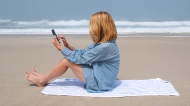 Atlantik Okyanusu sahilinde akıllı telefonunu kullanan genç bir kadın havluda oturuyor. Ekrana doğru el sallıyor ya da el sallıyor, muhtemelen bir video görüşmesinde, dalgalı bir sahil anının tadını çıkarıyor.