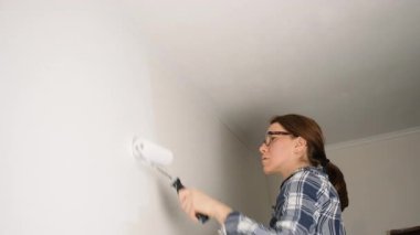 Tavana yakın bir duvarı boyamak için silindir kullanan genç bir kadın. Detaylı bir ev geliştirme sürecinin parçası olarak beyaz boyayı dikkatlice uyguluyor.