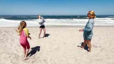 Anne ve iki çocuk sahilde birlikte oynuyorlar, aralarına su dolu bir balon atıyorlar. Neşeli oyun, parlak, güneşli bir gökyüzünün altında okyanus dalgaları tarafından gözler önüne seriliyor..