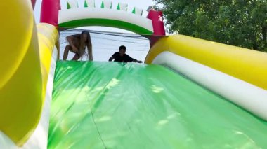 İki genç, bir açık hava etkinliğinde şişme bir kayaktan kayıyor. Gökyüzü ve yeşil ağaçların altında hareketle, kahkahalarla ve eğlenceyle dolu neşeli bir yaz anıydı.