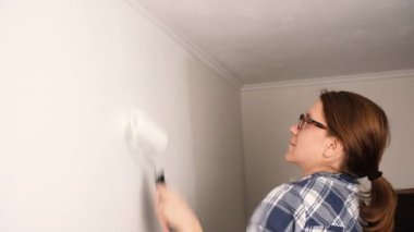 Beyaz paten fırçasıyla iç duvarı boyayan bir kadın. Duvarın üst kısımlarına odaklanmış durumda. Ev yenileme çalışmaları sırasında yeni bir kat boya sürüyor.