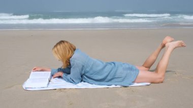 Genç bir kadın plaj havlusunda yüzüstü uzanmış Atlantik Okyanusu 'nun kıyısında kitap okuyor. Huzurlu ve güneşli bir günün tadını çıkarıyor. Arka planda dalgalar yavaşça çarpıyor.