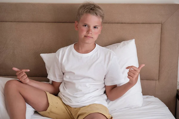 A cute young teenager sits in bed in a white t-shirt and points to his t-shirt. Mockup, copy space