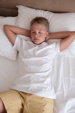 Calm young handsome teenager lies in bed in a white t-shirt in the bedroom at home. Rest, relaxation, good mood, lifestyle. Mockup, copy space