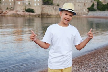 Sahil kenarında hasır şapkalı şık bir genç çocuk gülümsüyor ve beyaz bir tişörtü işaret ediyor. Model.
