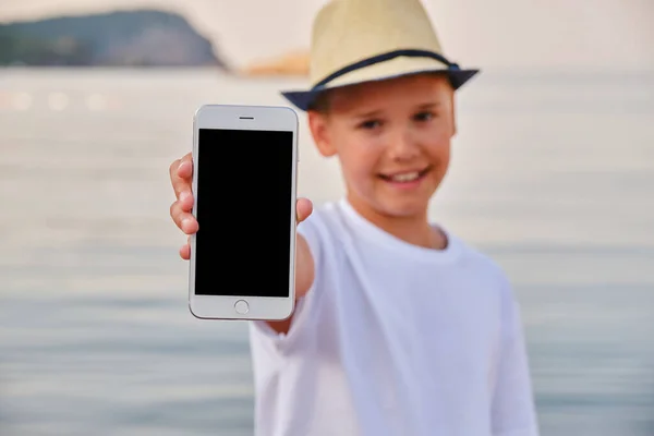 A stylish teenager in a straw hat on the seashore demonstrates the screen of a smartphone. Mobile app layout. Template for design.