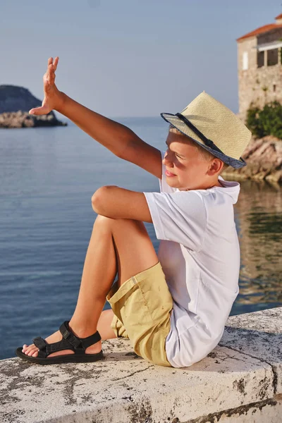 Mladý Chlapec Bílém Tričku Slaměném Klobouku Sedí Pobřeží Rukou Zakrývá — Stock fotografie