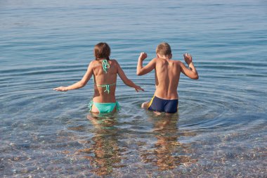 Erkek ve kız kardeşler soğuk, şeffaf denize girerler. Arkadan bak. Güneşli yaz günü, tatil günü