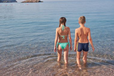 Erkek ve kız kardeşler berrak saydam denize girerler. Arkadan bak. Güneşli yaz günü, tatil günü