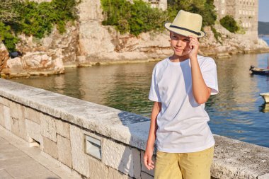 Deniz kenarında oturan beyaz tişörtlü ve hasır şapkalı bir gencin portresi. Modelleme. Yaz tatili ve seyahat kavramı.