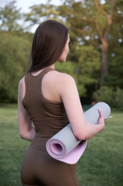 Yazın parkta dururken yoga minderi tutan genç bir kadın. Sağlıklı yaşam tarzı ve uzatma kavramı.