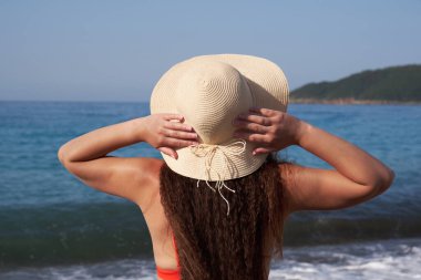 Hasır şapkalı genç bir kadın güneşli bir yaz gününde denize bakıyor..