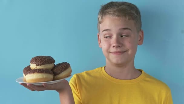 Щасливий Милий Підліток Вказує Повну Тарілку Шоколадних Пончиків Синьому Фоні — стокове відео