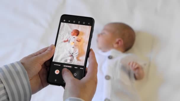 スマートフォンカメラで赤ちゃんの写真を撮る母親の近く ブロガーの母親の現代のオンライン生活 — ストック動画