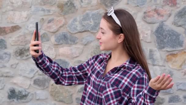 一位穿着格子衬衫的少女站在石墙边 一边在智能手机上流口水 一边录音 — 图库视频影像