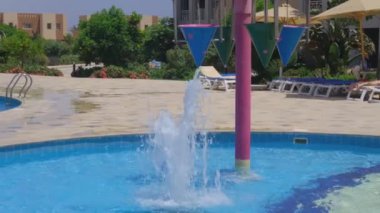 Havuz kenarında etkileşimli su özelliği, çocuklar renkli çöp kovalarıyla oyun alanı.