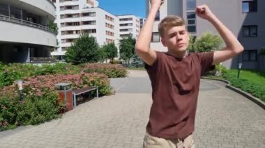 Genç çocuk, arka planda binalar ve yeşillikler olan bir yerleşim bölgesinde enerjik bir şekilde dans ediyor..