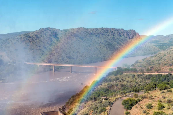 The single lane road bridge over the Orange River below the Gariep Dam. is visible behind a double rainbow