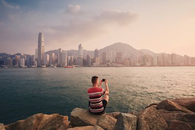 Gün batımında Hong Kong kentinin ufuk çizgisini fotoğraflayan adamın arka görüntüsü. Modern şehirde akıllı telefon kullanan bir turist