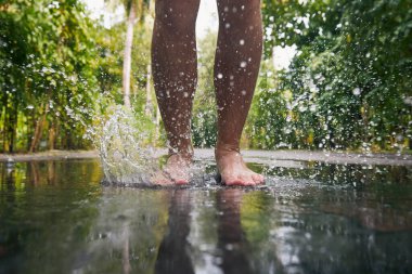 Man jumping with bare feet into puddle during rainy day. Close-up of legs and splashing water
