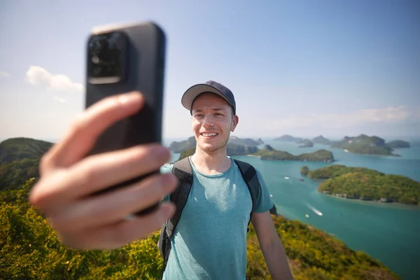 Smiling man with backpack taking selfie portrait on hill against group of tropical islands in sea. Ang Thong National Marine Park near Koh Samui in Thailand