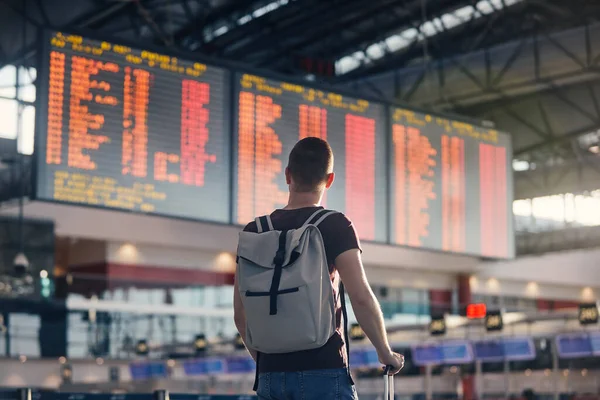 飛行機で旅行する バックパックとスーツケースを持った男が空港ターミナルを歩いて出発情報を見る — ストック写真