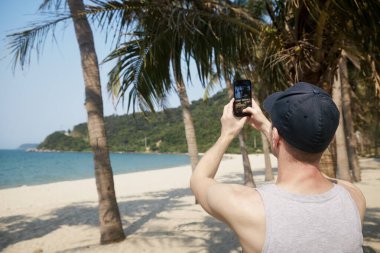 Cennet kumsalında akıllı telefon kullanan bir adam. Tropikal seyahat noktasında palmiye ağaçlarının fotoğrafını çekerken turistin arka görüntüsü.