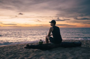 Hindistan cevizi içeceği ile tropik kumsalda oturan ve okyanusun üzerinde renkli günbatımının tadını çıkaran bir adam. Temalar yalnız ve buget seyahat