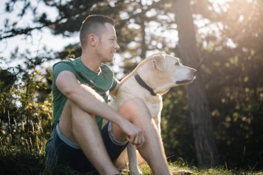 Ağacın altında çimlerde oturan sevimli köpekli bir adamın portresi. Güneşli yaz gününde doğada dinlenen Labrador Retriever 'ın mutlu sahibi.