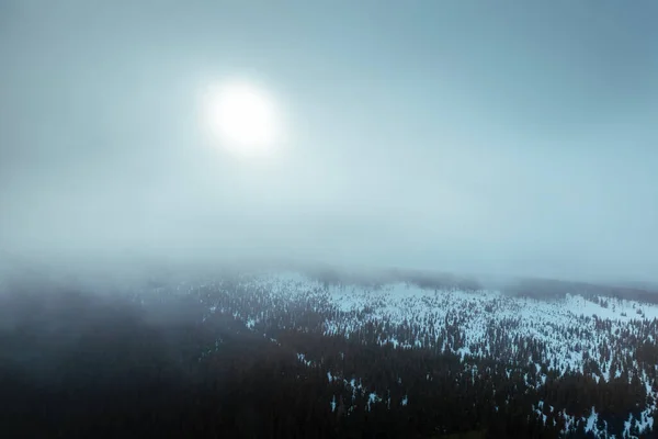 Aerial view of snowy mountain range in cloud with sun shining through during frosty day. Moody winter scenery