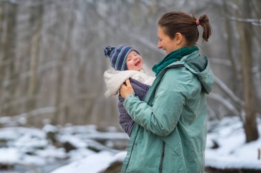 Neşeli bir anne, gülen bebeğini karlı bir ormanda tutuyor, sakin bir kış ortamında sevgi ve aile bağını gösteriyor, şefkati, mutluluğu ve mevsimin açık hava güzelliğini vurguluyor..