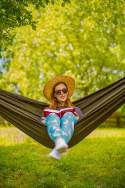 Güneş gözlüğü ve güneş gözlüğü takan bir kadının sakin bir sahnesi. Sıcak güneş ışığı altında bir hamakta dinleniyor. Canlı yeşil manzarayla çevrili bir kitap okuyor..