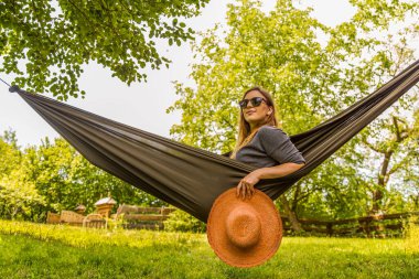 Kendine güvenen bir kadın elinde hasır şapkayla hamakta dinlenirken dışarıda eğleniyor. Sakin bahçe dekorasyonu sergiler yemyeşil ve canlı yaz titreşimleri, sükunet için mükemmel.