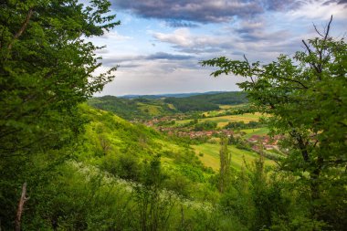 Uzakta bir köy ile yemyeşil manzara, yuvarlanan tepeler ile çevrili.