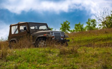 Engebeli bir off-road aracı mavi gökyüzünün altında yemyeşil bir yamaçta park etmiş..