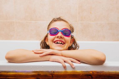 Gözlüklü çocuk küvetin kenarına yaslanıyor, mutluluk ve neşe saçıyor.