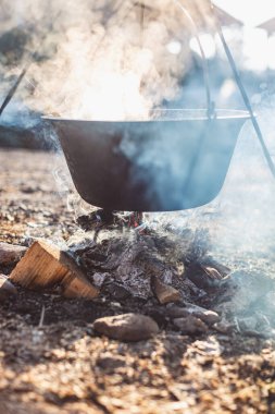 Duman ve ahşap odunlarla çevrili bir kamp ateşinin üzerinde dumanı tüten bir tencere asılı duruyor..