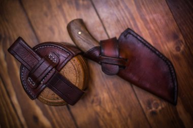 Ahşap tahtaların üzerinde deri aksesuarların yanında duran el yapımı deri bir bıçak kılıfı..