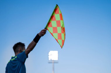 Futbol maçında hakemin bayrağı göndere çekildi. Arka planda mavi gökyüzü ve lambalar var..